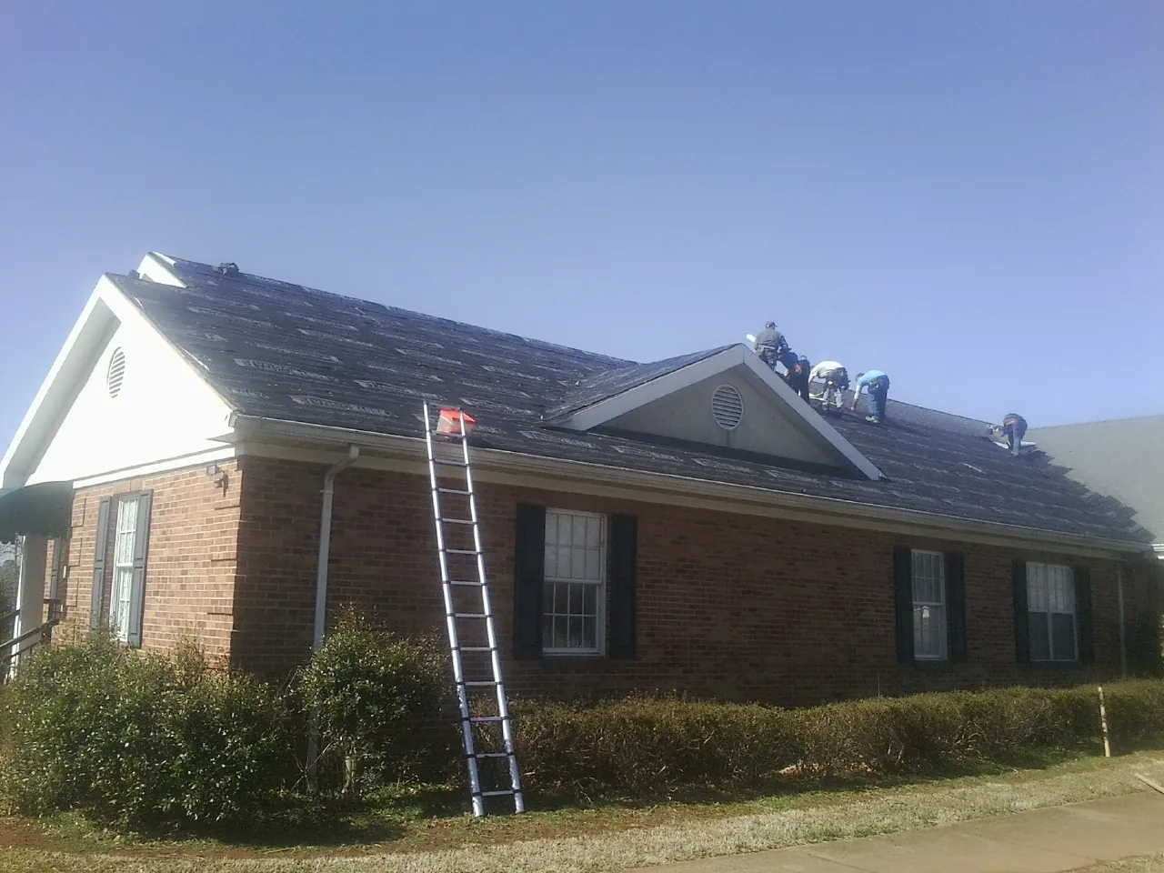 Crew tearing shingles off a brick ranch roof with a ladder set — Emergency Roof Repair work by our crew in Williamsburg